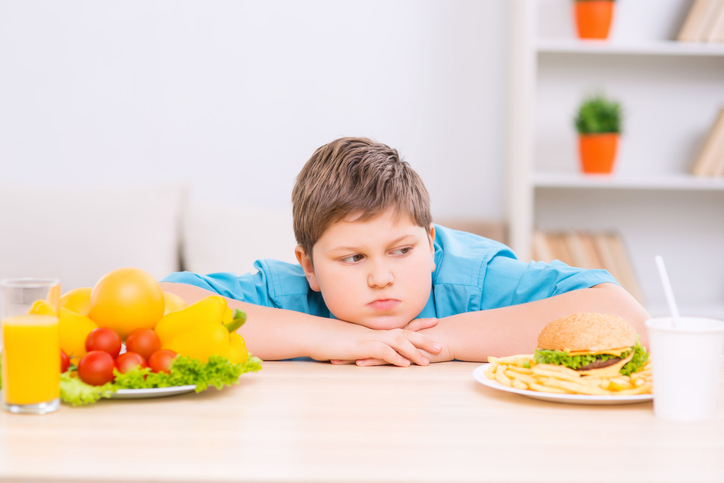 健康的な食べ物と不健康な食べ物の2つの皿の間に太った子供がいる。