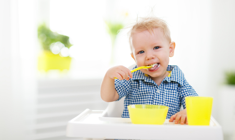 ベビーシートに座り、自分で食事をする小さな子供。 黄色いスプーン、カップ、ボウル。