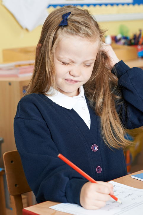 頭をかゆがり、髪にシラミがいるのだろう、金髪の少女が学校で座りながら書いている。